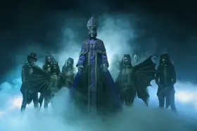 Le **chanteur** du groupe Ghost, vêtu d'une soutane violette, est entouré de musiciens masqués en squelettes, dans une atmosphère brumeuse et sombre.