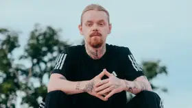 Mężczyzna z rudą brodą i blond włosami, ubrany w czarną koszulkę z trzema paskami. Na szyi i rękach widoczne tatuaże, w tym klucz i napis "kuqe 2115".