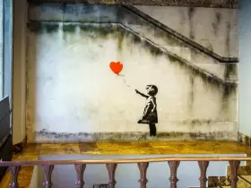 Banksy dziewczynka z balonikiem, unoszącym się na tle schodów i zniszczonej ściany.