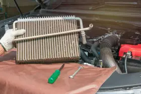 Gloved hand holds car AC evaporator. Tools and engine parts visible. Learn how to make AC colder in car by fixing this component.