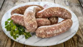 Kilka surowych kiełbasek białych parzonych ułożonych na marmurowej desce, ozdobione gałązką zielonych liści.