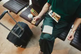Osoba w zielonej koszulce siedzi na lotnisku z walizką i kawą, jedząc kanapkę. Czy można zabrać do samolotu kanapki? Tak, jak widać!