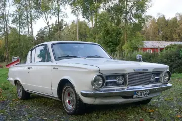 Vit Plymouth Barracuda från 1960-talet parkerad på en gräsmatta med träd och en röd byggnad i bakgrunden.