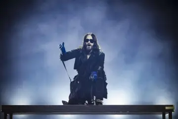 Jared Leto w niebieskich rękawiczkach, w okularach i długim płaszczu, klęczy na scenie. Jego zespół pewnie czeka na jego znak.