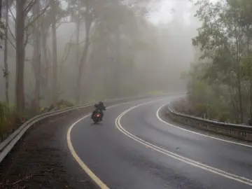 Motocyklista pokonuje zakręt na mokrej drodze, gdzie grozi mu aquaplaning. Mgła otula las, tworząc tajemniczą atmosferę.