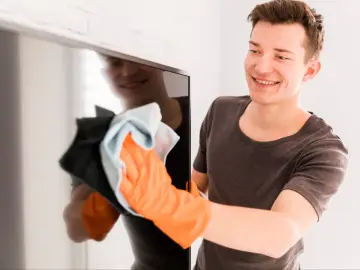 Uśmiechnięty mężczyzna w pomarańczowych rękawiczkach czyści ekran telewizora, pokazując, jak wyczyścić monitor bez smug.