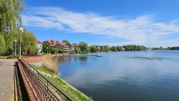 Spokojne jezioro zamkowe z fontanną i drewnianym pomostem, otoczone zielenią i zabudowaniami.
