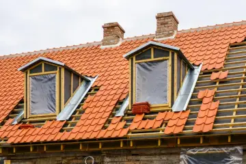 Trwa wymiana okna dachowego. Dach pokryty jest pomarańczową dachówką, a okna są tymczasowo zabezpieczone folią.