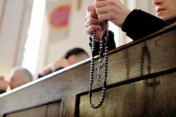 Dłonie trzymające różaniec podczas nabożeństwa. W tle widać innych wiernych, przygotowujących się na odpust.