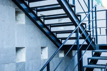 Metalowe schody z poręczą biegną wzdłuż szarej, kamiennej ściany z kwadratowymi wnękami. Murek zamiast balustrady tworzy ciekawy kontrast.