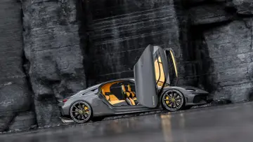 Szary Koenigsegg Gemera, czteromiejscny supersamochód z otwartymi drzwiami, prezentuje swoje żółte wnętrze na tle skalistej ściany.