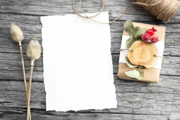 Jak zrobić zaproszenie na komunię w Wordzie? Pusta kartka, suszone maki i ozdobny prezent na drewnianym tle.