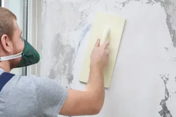 Mężczyzna w masce ochronnej nakłada tynk mozaikowy na ścianę za pomocą kielni.