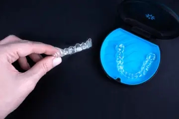 Dłoń trzyma przezroczysty aparat Invisalign. Obok, w niebieskim etui, leży kolejny.