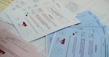 Karta do głosowania w wyborach samorządowych. Na stole leżą pakiety kart do głosowania, niektóre białe, inne niebieskie i różowe.