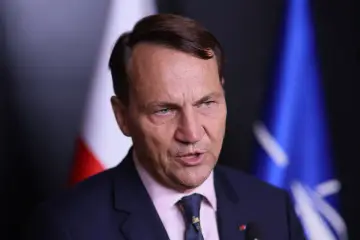 Minister spraw zagranicznych w garniturze, z polską flagą i flagą NATO w tle.