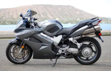 Szary motocykl Honda VFR 800 VTEC z widokiem na wodę i wzgórza w tle.