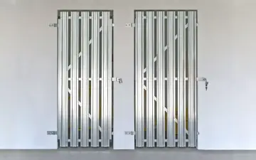 Dwa metalowe, harmonijkowe drzwi do piwnicy w bloku, zabezpieczone kłódkami.