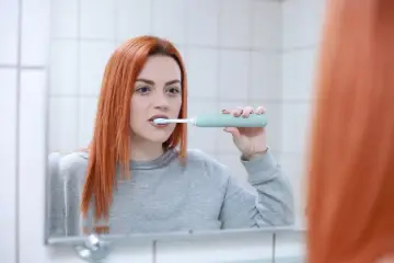 Rudowłosa kobieta myje zęby szczoteczką soniczną, zastanawiając się, czy szczoteczka soniczna usuwa kamień.