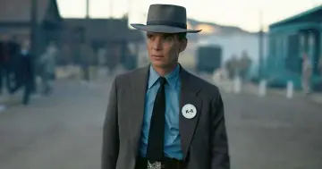 Julius Robert Oppenheimer w kapeluszu i garniturze, z odznaką K-6, w scenerii przypominającej miasteczko.