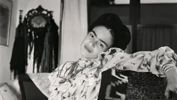 Frida Kahlo, le regard intense, pose dans une robe à volants. Derrière elle, un objet décoratif orné de franges, peut-être un indice sur son dernier tableau.
