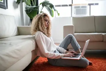 Kobieta z kręconymi włosami pracuje na laptopie na dywanie, opierając się o kanapę. Czy na macierzyńskim można pracować? Tak, zdalnie.