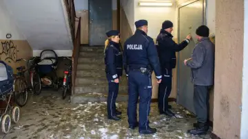Policja puka do drzwi, by sprawdzić, czy nadzór budowlany może wejść do mieszkania. Na klatce schodowej stoją wózki i rowery.