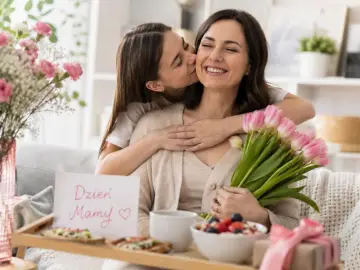 Córka całuje mamę w policzek, wręczając jej bukiet różowych tulipanów. Na tacce śniadaniowej kartka z napisem "Dzień Mamy".