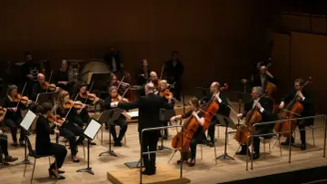 Orkiestra podczas koncertu. Dyrygent prowadzi muzyków grających na instrumentach smyczkowych.
