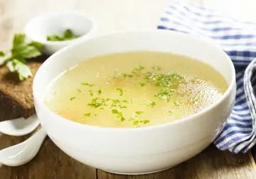 Aromaticzny rosół z perliczki, ozdobiony natką pietruszki, podany z kromką chleba i łyżeczkami.