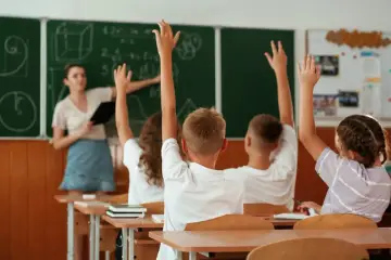 19 maja. Nauczycielka przy tablicy, uczniowie z podniesionymi rękami, gotowi do odpowiedzi.