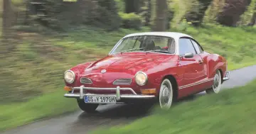 Czerwony Karmann Ghia z białym dachem pędzi po mokrej drodze wśród zieleni.