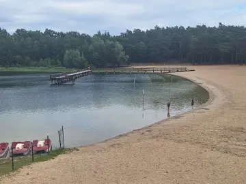 Drewniany pomost nad jeziorem Kamionki, piaszczysta plaża, łódki i dwie osoby kąpiące się w wodzie.
