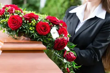 Wieniec z czerwonych róż na drewnianej trumnie, obok osoba w czarnym garniturze.