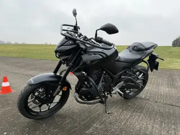 Czarny motocykl Yamaha MT-03 stoi na betonowej nawierzchni, gotowy do jazdy po zielonym polu.