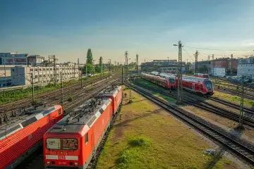 Czerwone lokomotywy i wagony stoją na torach. W tle budynki i drzewa. To miejsce, gdzie przygotowuje się kolej dużych prędkości do podróży.