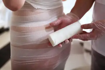 Osoba w rękawiczkach trzyma biały wałek, podczas gdy druga osoba owija ciało bandażem. Widoczne body wrapping efekty.