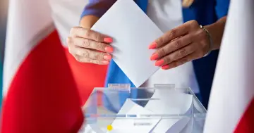 Kobieta z pomarańczowym lakierem na paznokciach wrzuca kartę do urny wyborczej. W tle polskie flagi, symbolizujące głosowanie za granicą.