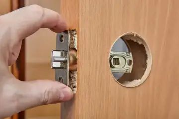 Dłoń montuje mechanizm zamka w drewnianych drzwiach. Dowiedz się, ile kosztuje wymiana zamka w drzwiach.