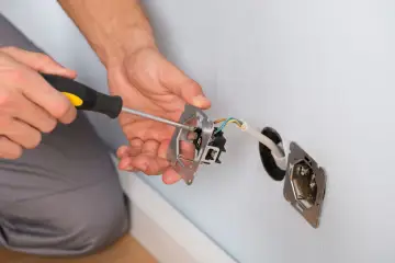Ręce majsterkowicza przykręcają śrubokrętem mechanizm gniazdka. Widać przewody elektryczne i puszkę instalacyjną.