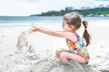 Dziewczynka w kolorowym stroju kąpielowym z okularami na nosie rzuca piasek, ciesząc się zabawami na plaży.