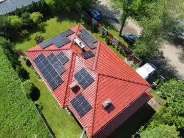 Dach z panelami fotowoltaicznymi, które stanowią dodatek energetyczny dla domu. Widać też samochody i zieleń.