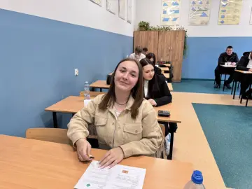 Uśmiechnięta uczennica w technikum, kiedy jest półmetek. W tle inni uczniowie przy ławkach, na ścianach tablice z matematyki.