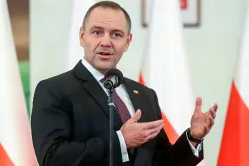 Premier Polski w garniturze, przemawiający przy mikrofonie na tle flag.