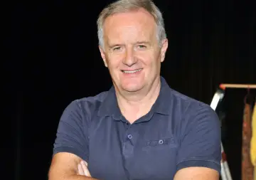 Aleksander Mikołajczak, uśmiechnięty mężczyzna w granatowej koszulce polo, z rękami skrzyżowanymi na piersi.