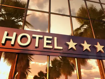Elegancki hotel z napisem "HOTEL" i czterema gwiazdkami. W szklanej fasadzie odbijają się palmy i zachodzące słońce.