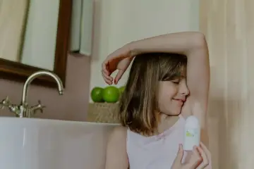 Uśmiechnięta dziewczynka zamyka oczy, aplikując dezodorant dla dzieci. W tle widać wannę i jabłka.
