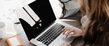 Kobieta siedzi przy laptopie, szukając informacji, jak sprawdzić status bezrobotnego. Obok stoi kubek kawy i pomarańczowy dysk zewnętrzny.