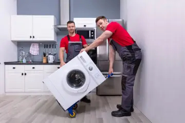 Dwóch mężczyzn w szarych ogrodniczkach i czerwonych koszulkach przewozi pralkę na wózku. Czy można przewozić pralkę na leżąco? Zapytaj fachowców.