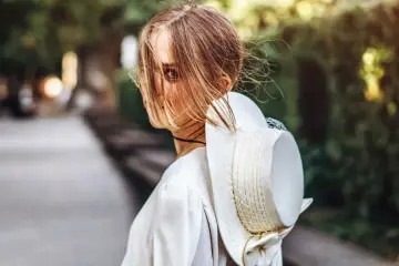 Kobieta z pięknymi bronde włosami, częściowo zasłoniętymi przez wiatr, odwraca głowę, trzymając elegancki kapelusz.